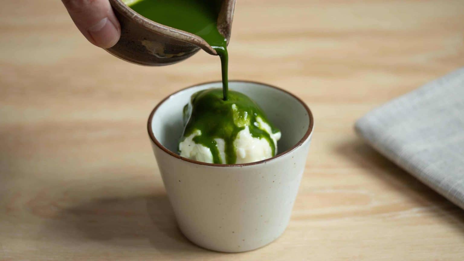 Green matcha sauce pours onto a scoop of vanilla ice cream in a white cup.