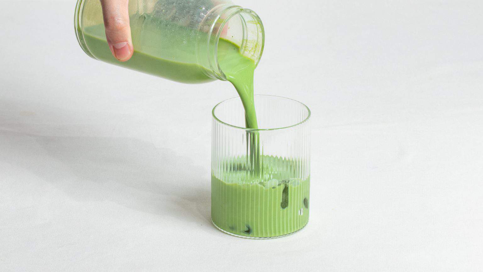 Pouring bright green matcha latte into a ribbed glass.
