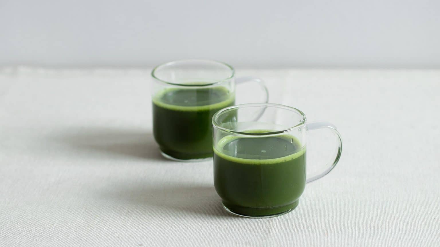 Two clear glass mugs filled with bright green matcha on a light fabric surface.