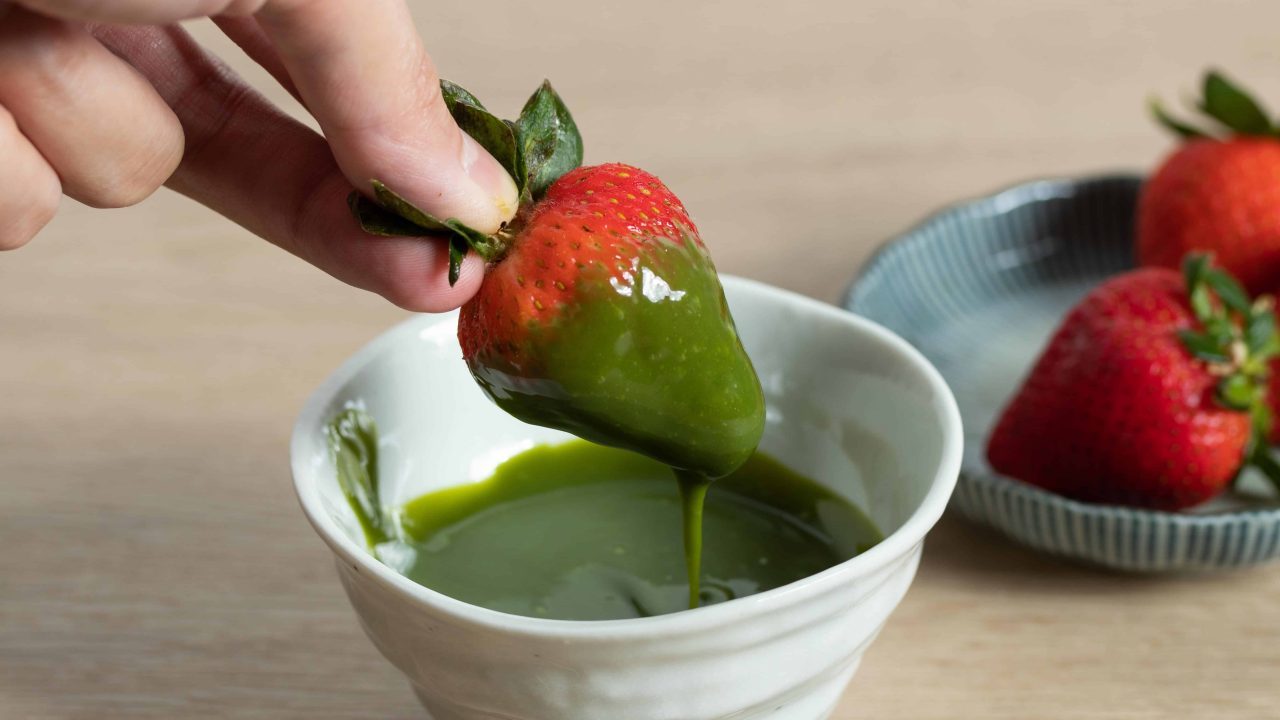 Strawberry being dipped into bright green matcha spread in a small white cup.
