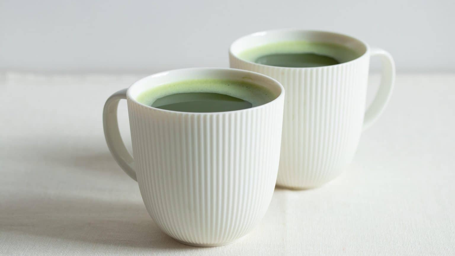 Two white ribbed ceramic cups filled with green matcha on a pale surface.