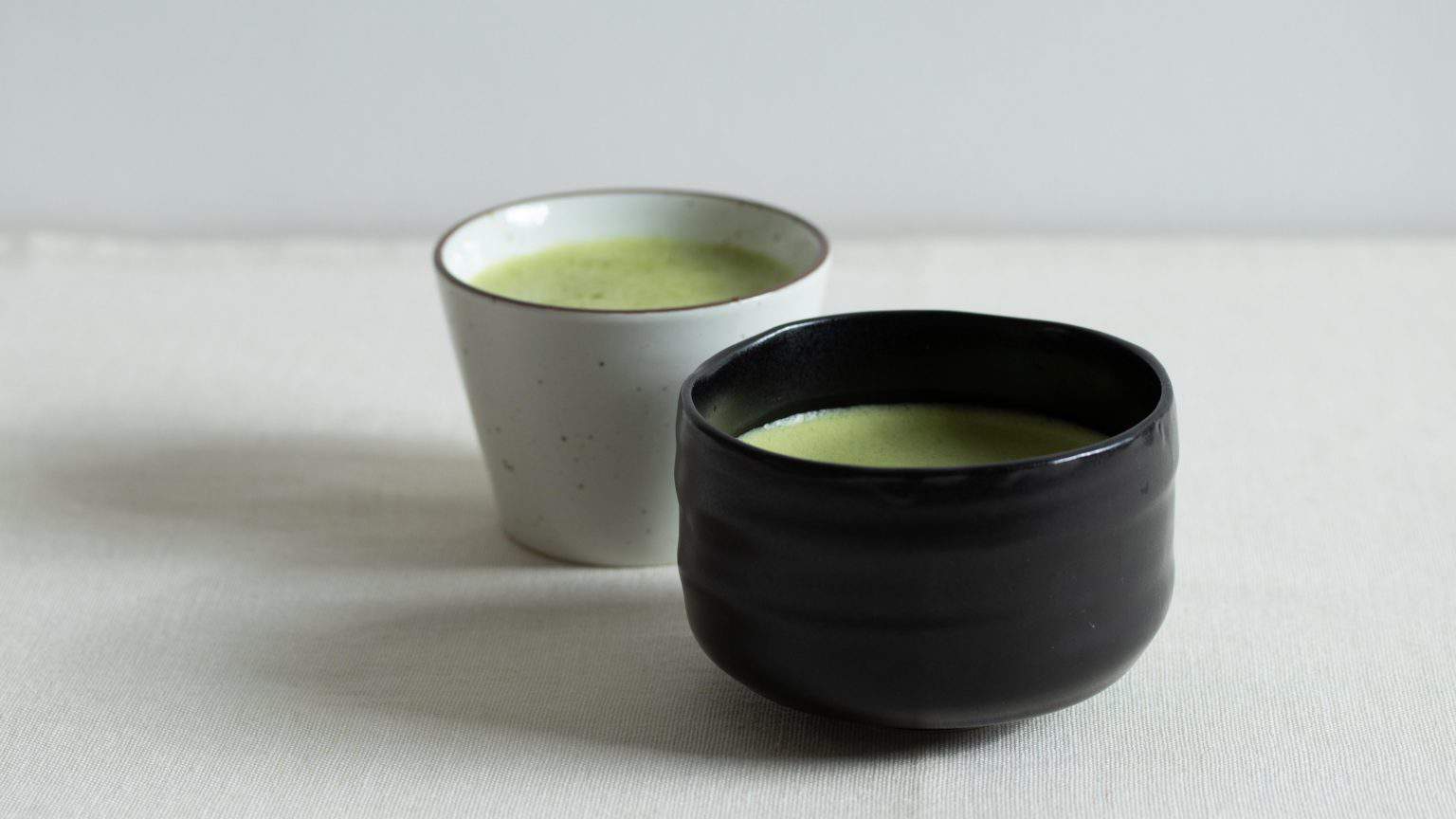 Two bowls of matcha tea on a light surface with a black ceramic bowl in front and a white cup behind