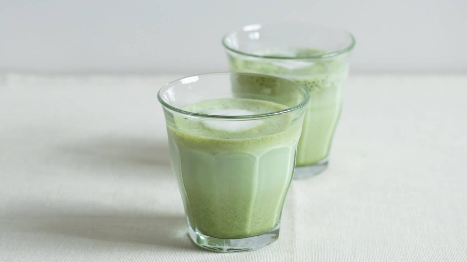 Two glasses of frothy green matcha latte on a light surface