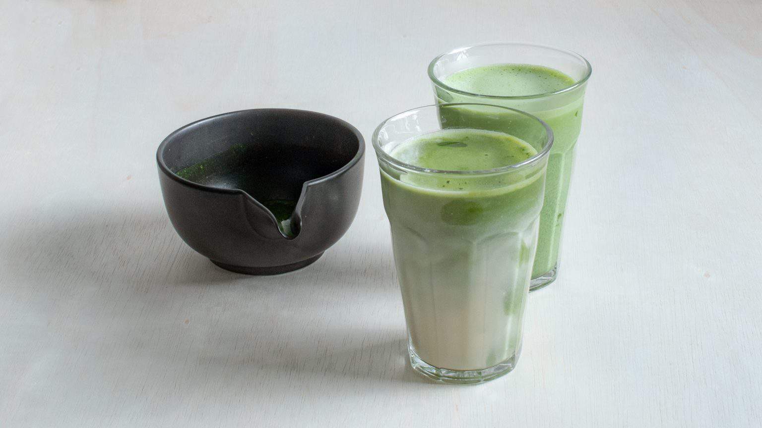 Two glasses of matcha oat milk latte on a white table with a bowl of matcha paste