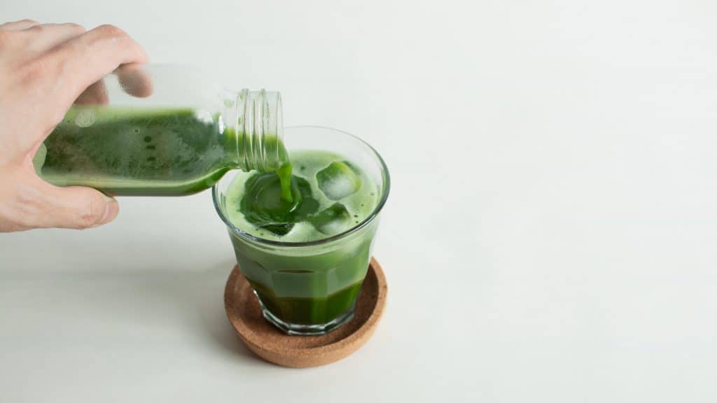 Hand pours vibrant green matcha from a bottle into a glass with ice on a cork coaster.