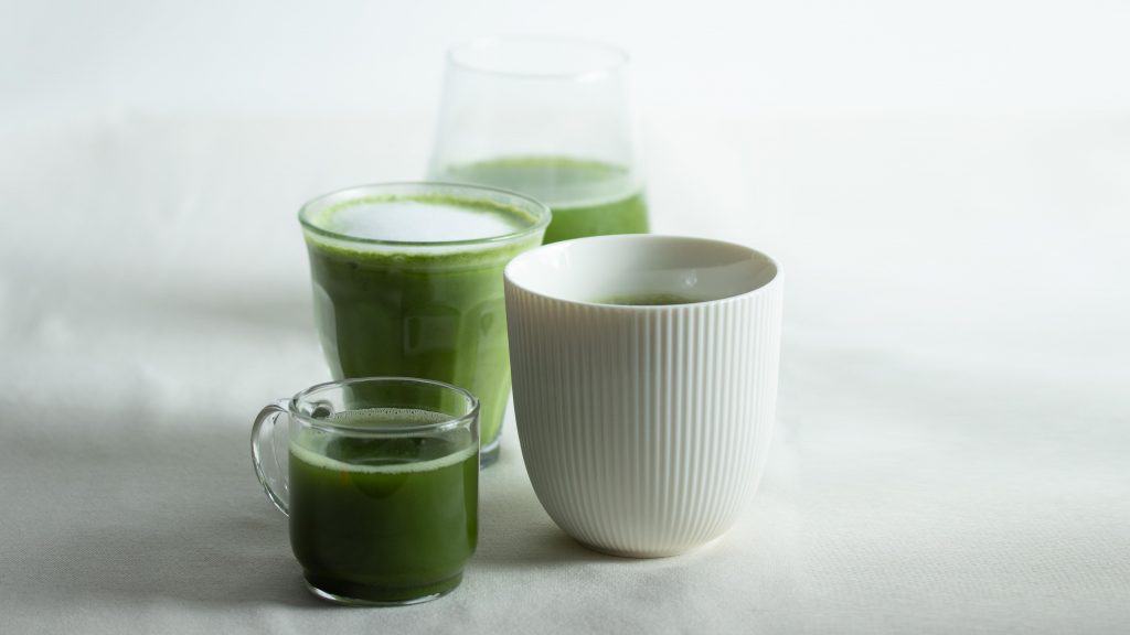 Three matcha drinks on a white surface including a ribbed white mug and two glass cups