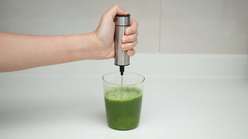 Hand using a stainless steel handheld whisk to blend bright green matcha in a glass