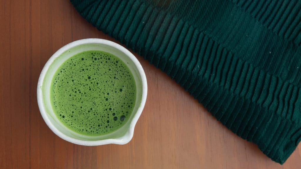 Bright green matcha tea in a white ceramic cup on a wooden table beside a dark green knit blanket.