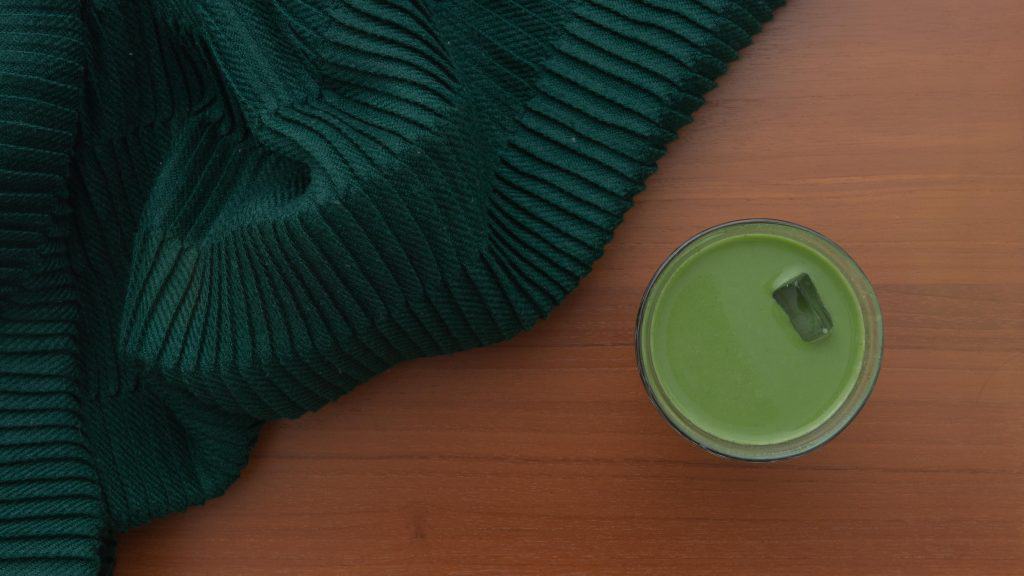 Glass cup of bright green matcha on a wooden table with a dark green knit fabric beside it