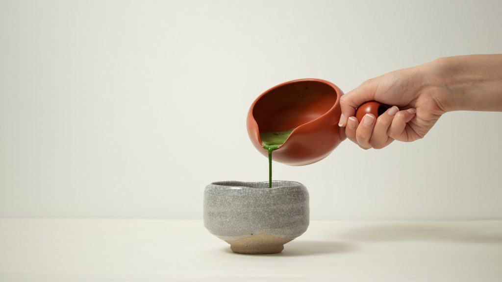 Hand pours bright green matcha from a brown pitcher into a gray ceramic cup