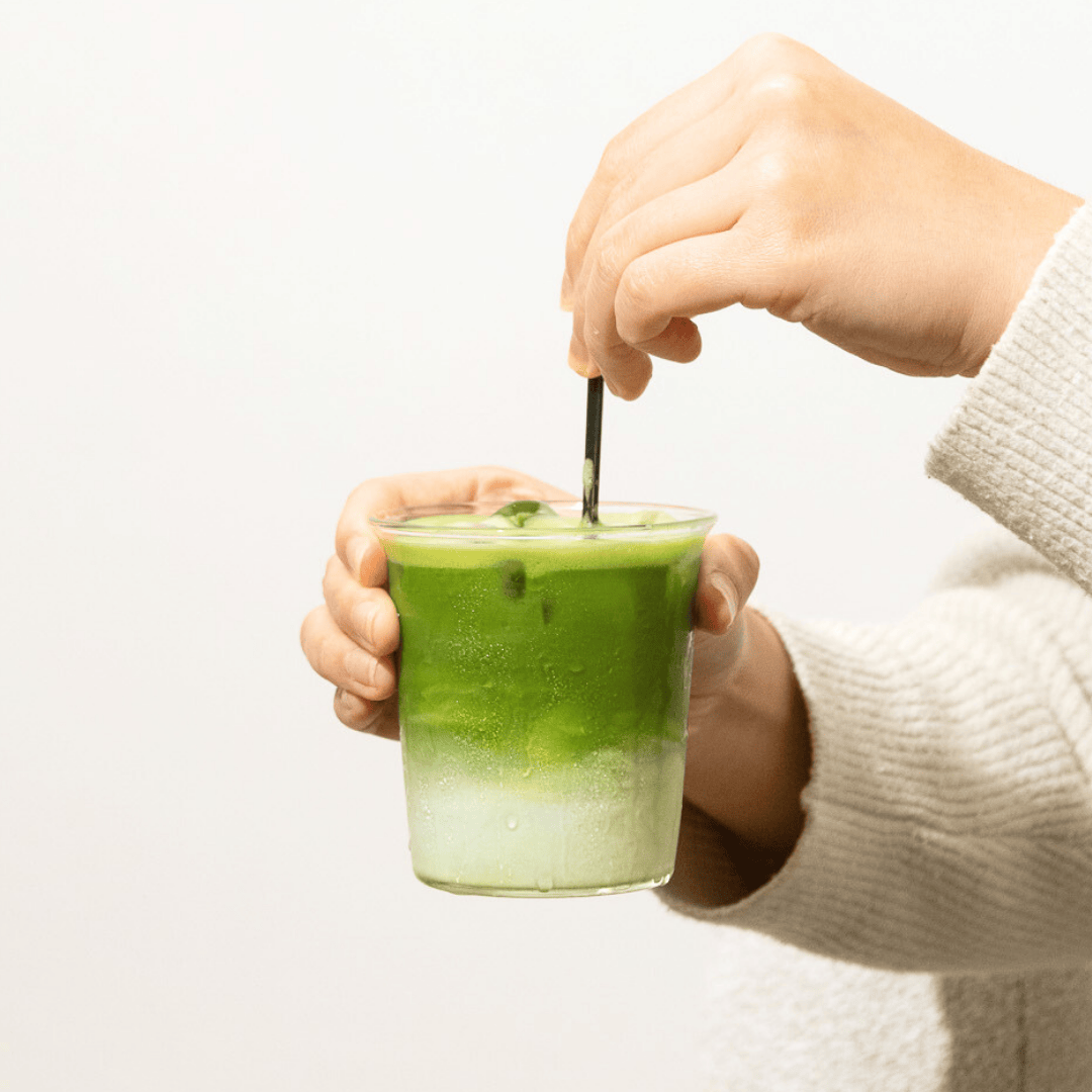 Green iced matcha latte in a glass stirred with a black straw by a person wearing a cream sweater