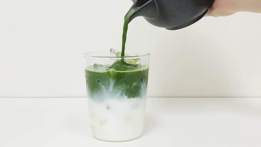 Pouring green matcha into a glass of milk with ice to make a latte